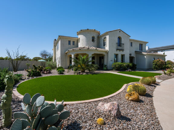 A Mediterranean-style home with a pop of green synthetic grass front lawn surrounded by desert plants and xeriscaping, all set against a bright blue sky.