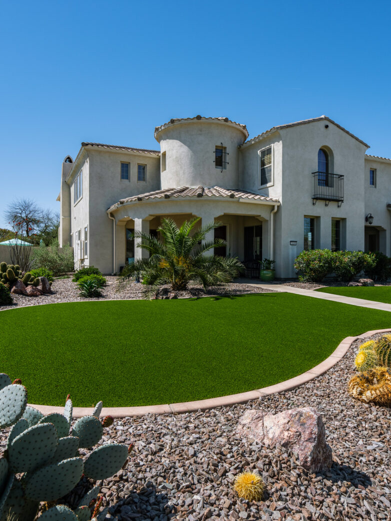 A Mediterranean-style home with a pop of green synthetic grass front lawn surrounded by desert plants and xeriscaping, all set against a bright blue sky.