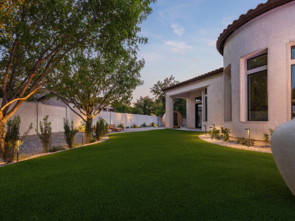 The front and side of a Mediterranean-style hom with an expansive swathe of dark, perfectly green synthetic grass illuminated by outdoor lights.