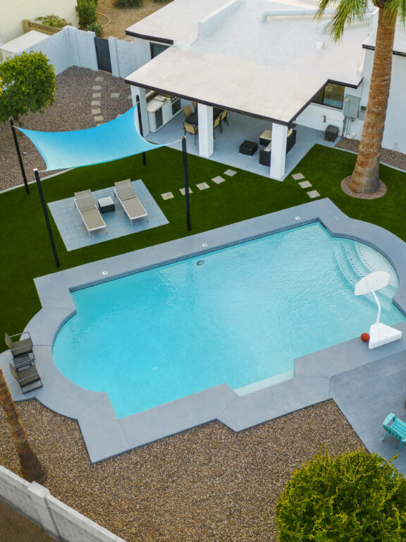 Aerial view of a backyard with a free-form swimming pool, artificial turf lawn, palm trees, shaded lounge area, and a covered patio beside a white modern home.