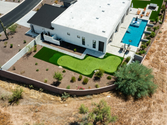 Aerial view of a modern desert home with a long rectangular swimming pool, paver patio, and a geometric artificial turf putting green within gravel landscaping and a fenced backyard.