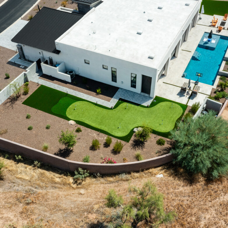 Aerial view of a modern desert home with a long rectangular swimming pool, paver patio, and a geometric artificial turf putting green within gravel landscaping and a fenced backyard.