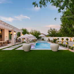 Backyard patio at dusk with artificial turf lawn, a rectangular swimming pool, shaded seating area, pergola with hanging lights, and desert landscaping along a white privacy wall.