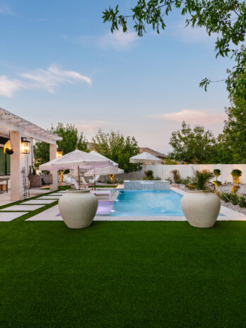 Backyard patio at dusk with artificial turf lawn, a rectangular swimming pool, shaded seating area, pergola with hanging lights, and desert landscaping along a white privacy wall.