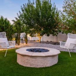 Backyard seating area with a circular fire pit set in artificial turf, two white lounge chairs, and surrounding desert landscaping with trees and gravel.