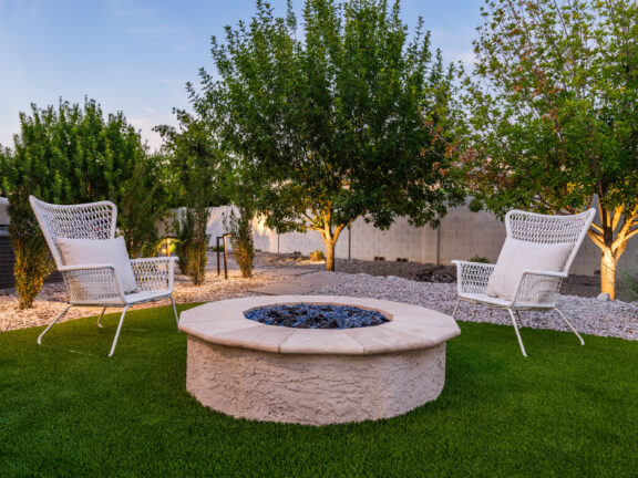Backyard seating area with a circular fire pit set in artificial turf, two white lounge chairs, and surrounding desert landscaping with trees and gravel.