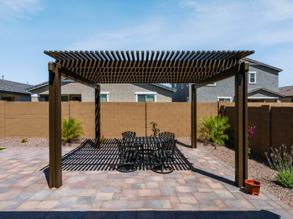 Backyard pergola with a slatted wood roof, paver patio flooring, and an outdoor dining table set against tan block walls and desert landscaping.