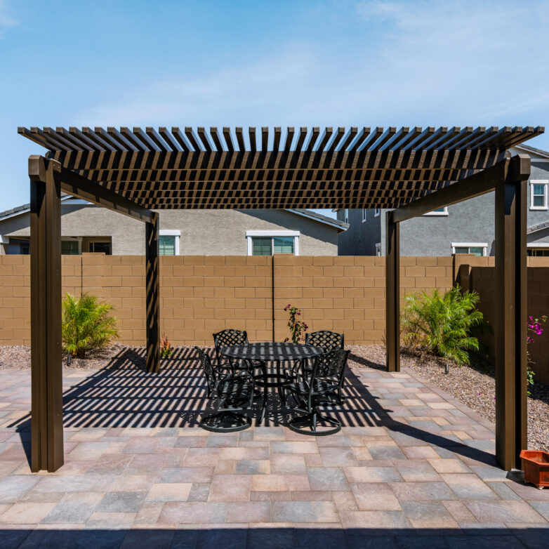 Backyard pergola with a slatted wood roof, paver patio flooring, and an outdoor dining table set against tan block walls and desert landscaping.