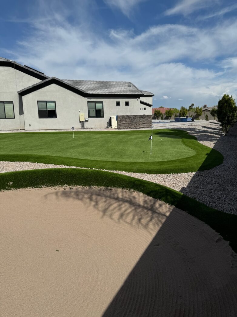 A backyard putting green with multiple golf holes, bordered by artificial turf and gravel, set behind a modern single-story house, with a sand trap in the foreground.