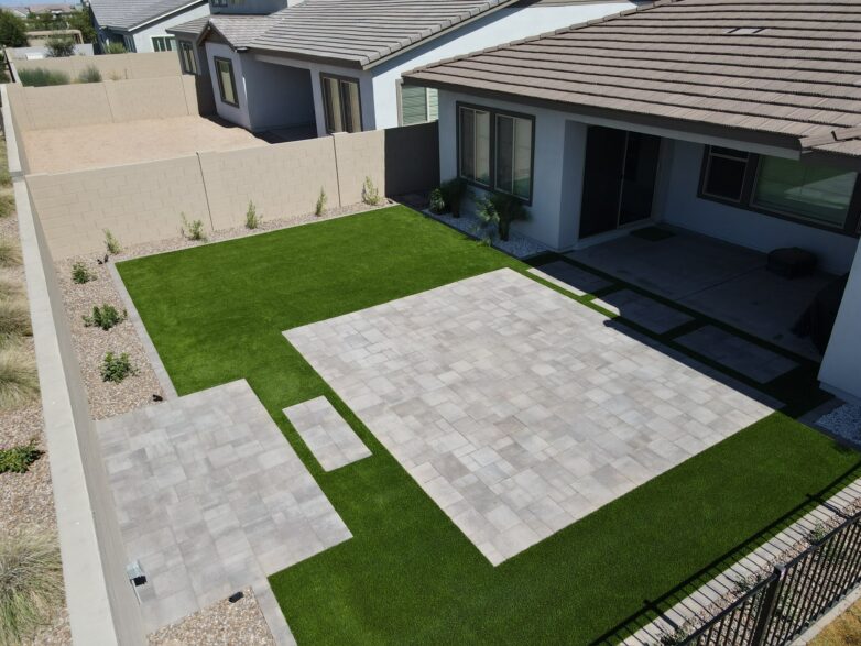 Backyard patio with light gray pavers, artificial turf lawn, and clean desert landscaping beside a modern stucco home.