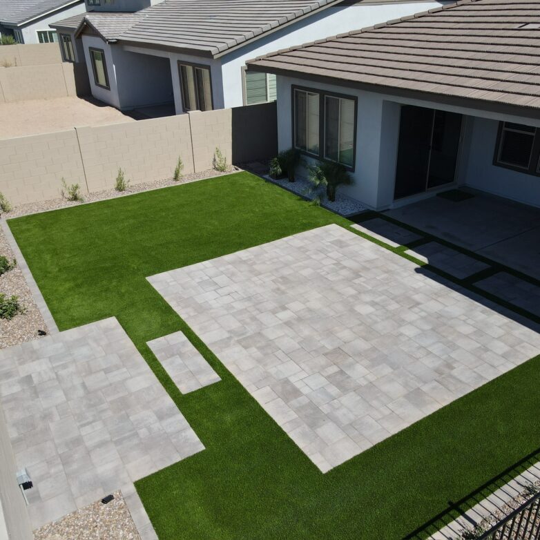 Backyard patio with light gray pavers, artificial turf lawn, and clean desert landscaping beside a modern stucco home.